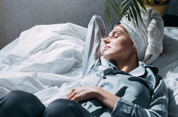 Een vrouw in pyjama en met een handdoek op haar hoofd ligt in bed op haar vanuit het raam, de ochtendzon schijnt, ze is ontspannen en sereen