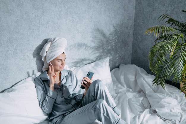 Een vrouw in pyjama en met een handdoek op haar hoofd zit op een bed in de ochtendzon en luistert naar muziek op een koptelefoon via de applicatie in een mobiele telefoon