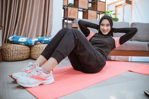 Een vrouw in sportkleding met hijab glimlacht terwijl ze sit-ups doet en haar buikspieren traint tijdens activiteiten thuis