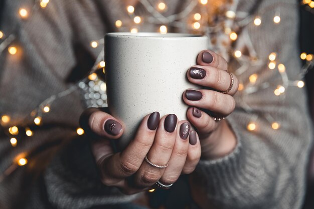 Een vrouw in warme kleding van neutrale tinten met een kerstslinger op haar knieën verwarmt haar handen aan een wit geëmailleerde mok met warme chocolademelk en marshmallows. De sfeer van gezelligheid en kerst.