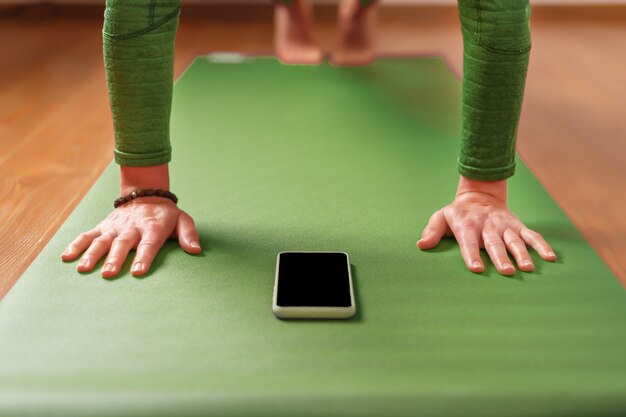 Een vrouw kijkt naar yogalessen op haar smartphone terwijl ze op een groene oefenmat zit. Het concept van training in de woonkamer van het huis. Technologie en sport