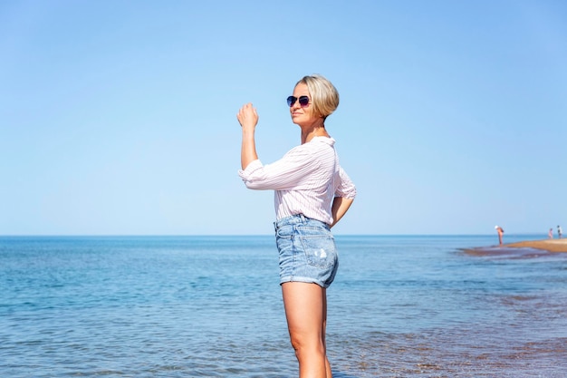 Een vrouw loopt op een zonnige dag langs de kust Een schoonheid met een stijlvol kapsel in een spijkerbroek en een licht gestreept shirt staat in het water Plezier van rust in het resort en reizen