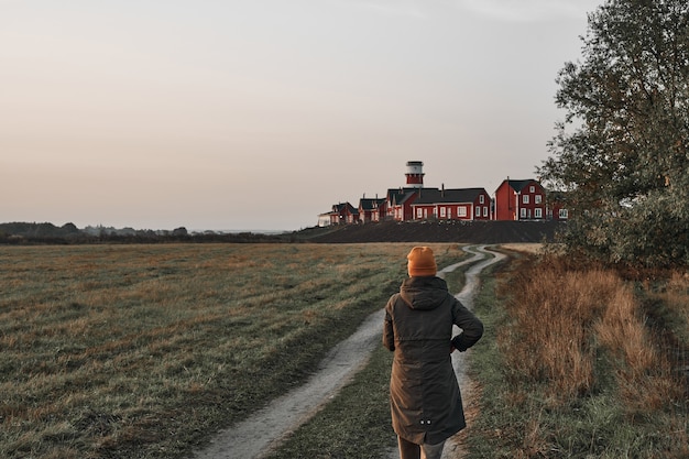 Een vrouw loopt over een landweg, een rood-witte vuurtoren aan het einde van de weg. Reis, dageraad.