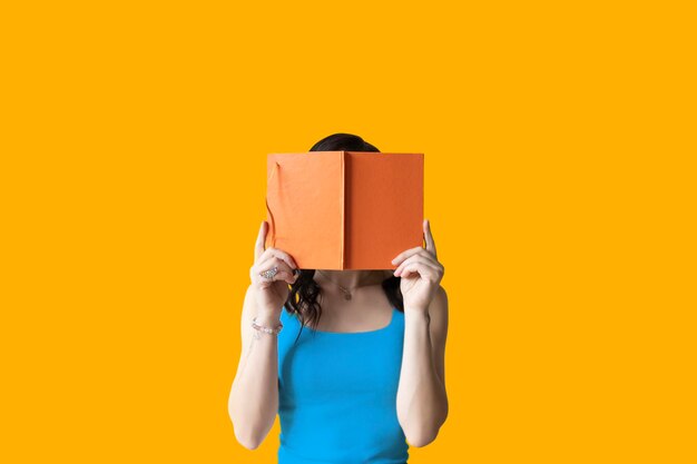 Foto een vrouw met donker haar bedekt haar gezicht met een oranje boek op een gele achtergrond