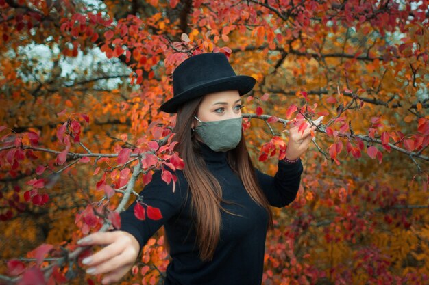 een vrouw met een beschermend masker is gekleed in een zwart jasje en hoed tegen een herfstpark