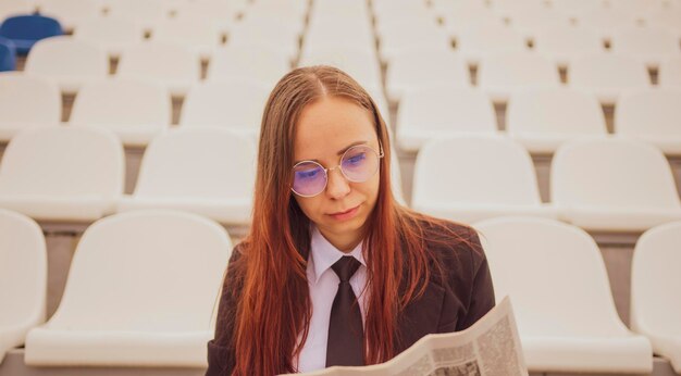 Een vrouw met een bril en een pak leest een krant terwijl ze in het stadion zit