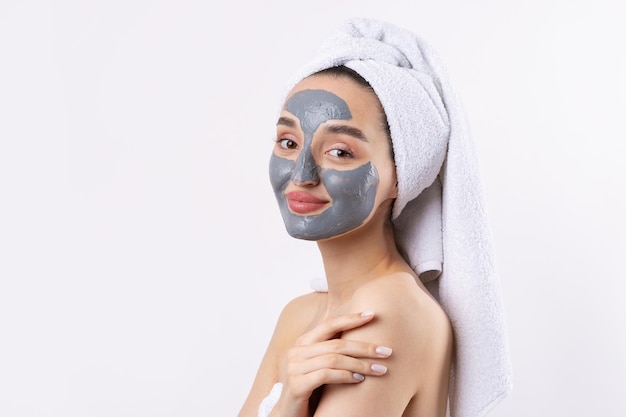 Een vrouw met een grijs cosmetisch masker van klei in een witte handdoek op haar hoofd op een witte achtergrond