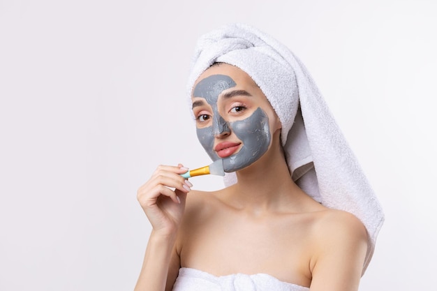 Een vrouw met een grijs cosmetisch masker van klei in een witte handdoek op haar hoofd op een witte achtergrond