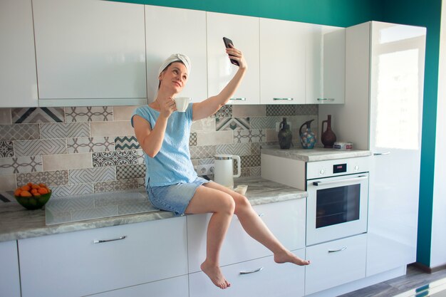 Een vrouw met een handdoek over haar hoofd zit na het douchen in de keuken met een kopje koffie en fotografeert zichzelf op een smartphone