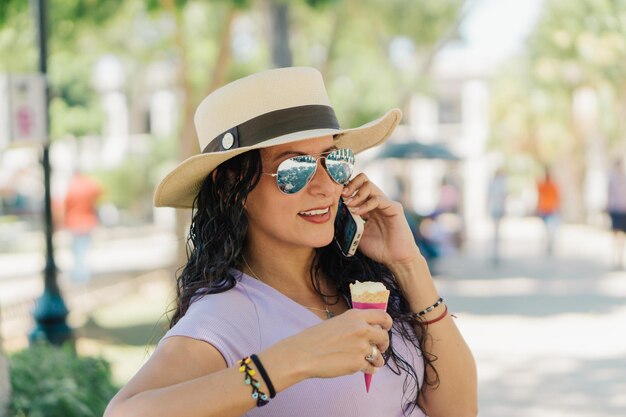 Een vrouw met een hoed en een zonnebril praat aan de telefoon terwijl ze een ijshoorntje vasthoudt.