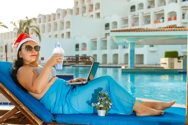 Een vrouw met een kerstmuts en zonnebril en een zwempak in een ligstoel met een laptop bij het zwembad van het hotel tijdens de kerstvakantie.