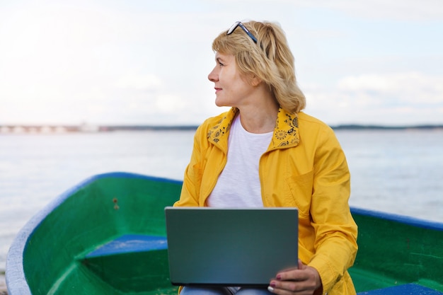 Een vrouw met een laptop aan het meer in de zomer. Een vrouw van middelbare leeftijd loopt langs het strand. Ze kijkt naar sociale media. Afstandswerk.