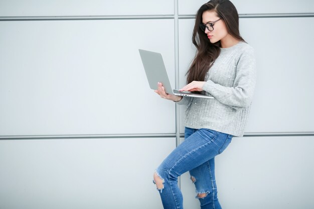 Een vrouw met een laptop in haar handen draagt een bril voor visie staat in de buurt van een grijze muur