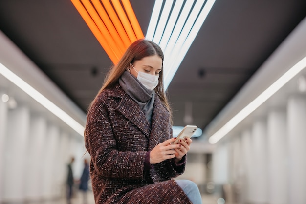 Een vrouw met een medisch gezichtsmasker zit in het midden van het metroplatform met een smartphone en leest het nieuws. Een meisje met lang haar met een chirurgisch masker houdt sociale afstand in de metro.