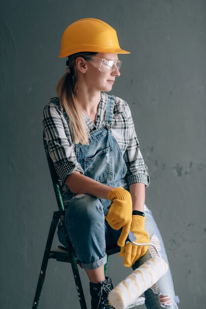 Een vrouw met een sterke wil in een bouwhelm, wanten, bril en overalls is thuis bezig met reparatie- en bouwwerkzaamheden. concept van een sterke en onafhankelijke vrouw