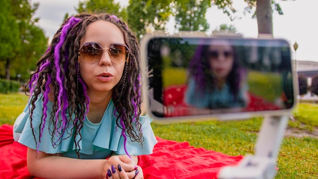 Een vrouw met een stijlvol kapsel ligt op een deken in het park en neemt een videoblog op met haar smartphone Een vrouwelijke blogger maakt op een zonnige zomerdag een video op een camera op een statief