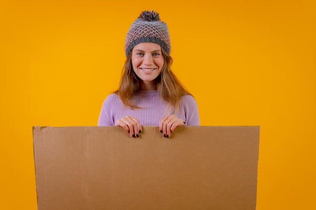Een vrouw met een wollen muts met een kartonnen bord op een gele achtergrond
