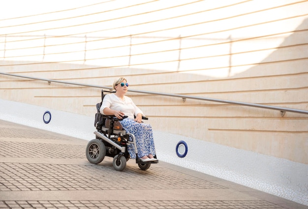 Een vrouw met een zonnebril die in een elektrische rolstoel zit en een stedelijke oprit afdaalt Concept van een voor gehandicapten toegankelijke stad