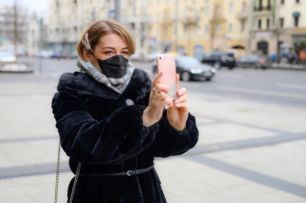 Een vrouw met een zwart medisch masker maakt een selfiefoto op een buitenwinter in de stad