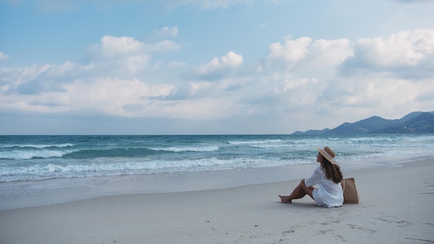 Een vrouw met hoed en tas zittend op het strand met blauwe hemelachtergrond