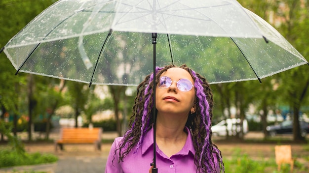 Een vrouw met paraplu in de hand in de regen Meisje met bril onder een paraplu tegen de achtergrond van een stedelijk landschap Vrouw met dreadlocks staat in een park met een paraplu