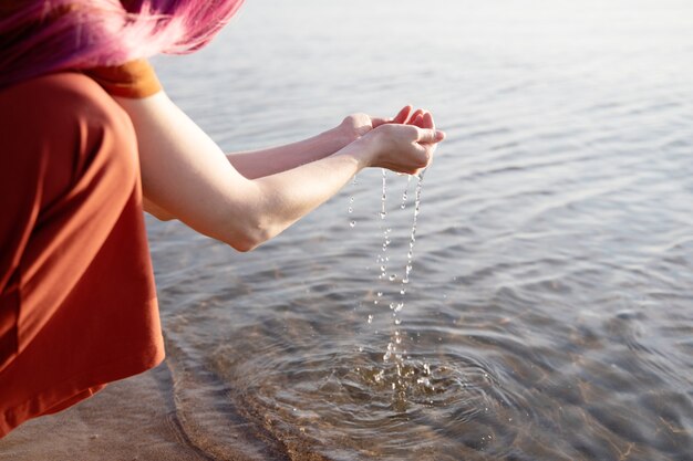 een vrouw met roze haar en oranje kleren pakt water uit de zee in haar handpalmen