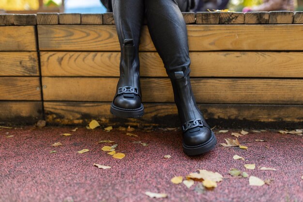 Een vrouw met zwarte laarzen zit op een bankje met bladeren op de grond.