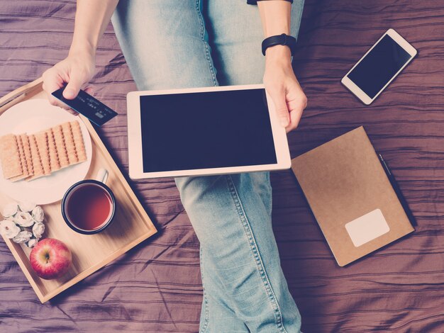 Een vrouw op bed zitten, thee drinken en kijken naar tablet, online winkelen.