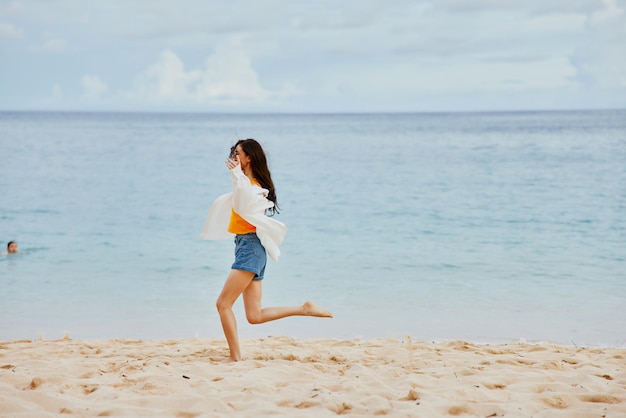 Een vrouw rent langs het strand in zomerkleren op het zand in een geel T-shirt en een spijkerbroek wit overhemd wapperend haar uitzicht op de oceaan