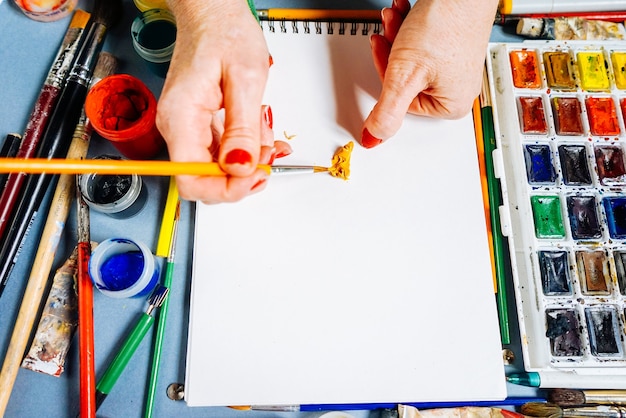 Een vrouw schildert een tekening op wit papier met een penseel, daarnaast is er een veelkleurige gouache en aquarellen