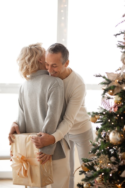 een vrouw staat bij een kerstboom en verbergt een cadeau voor haar man achter haar rug