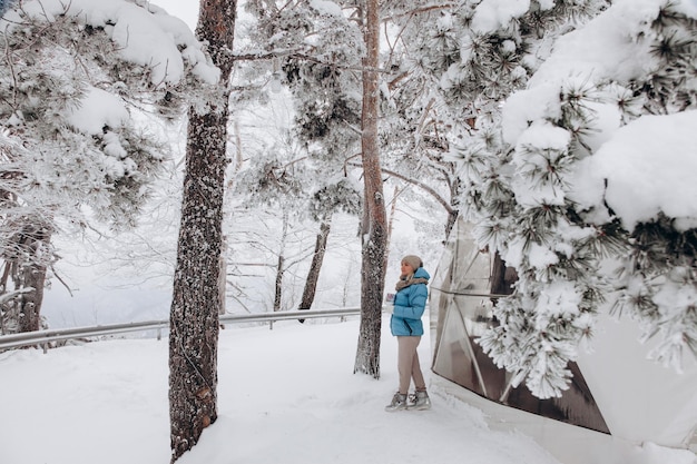 Een vrouw staat en kijkt naar de natuur met een thermocup in haar handen naast een koepeltent in glamping in het winterbos