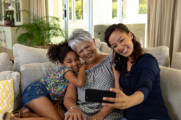 Een vrouw van gemengd ras thuis in de woonkamer, zittend op de bank met haar senior moeder en haar dochter en een smartphone gebruikend om een selfie van hen samen te maken, allemaal glimlachend aan de telefoon