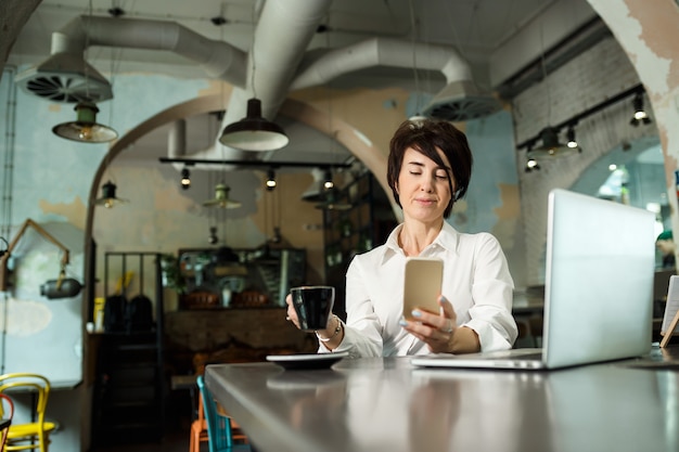 Een vrouw van middelbare leeftijd zit in een café, drinkt koffie en werkt achter een computer. vrouw met een telefoon