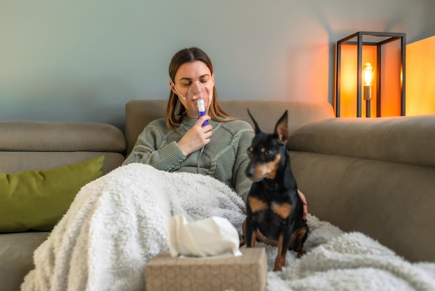 Een vrouw van middelbare leeftijd zit op een bank en inhaleert door een vernevelaar naast een hond