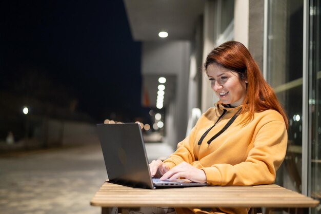 Een vrouw werkt laat in de avond op afstand achter een laptop in een zomercafé Een lachend meisje studeert zittend in een verlaten straat aan een houten tafel