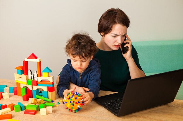 Een vrouw werkt thuis met een kind. de moeder zit achter een laptop terwijl het kind speelt en maakt een nepcoronavirus, en bouwt ook een huis van kubussen voor het gezin.