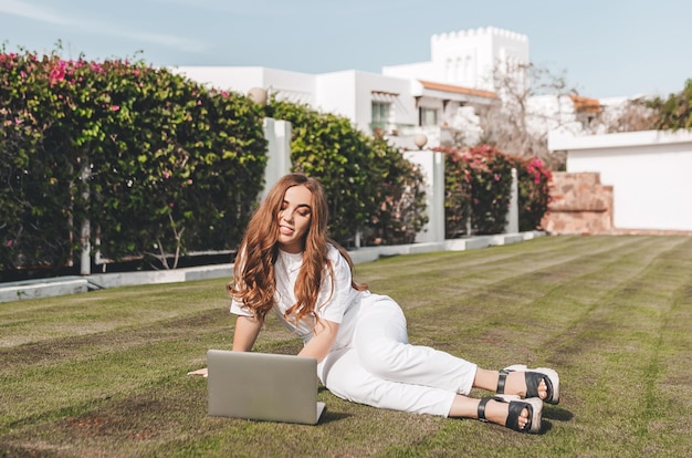 Een vrouw werkt thuis op afstand met een laptop op het grasveld Een meisje met een online beroep werkt overal ter wereld Vrolijk meisje op het gras in een warm klimaat