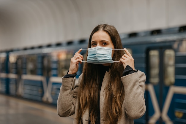 Een vrouw zet een medisch masker op haar gezicht om verspreiding van het coronavirus in een metrostation te voorkomen. Een meisje met lang haar in een jas wacht op een trein op een metroplatform.