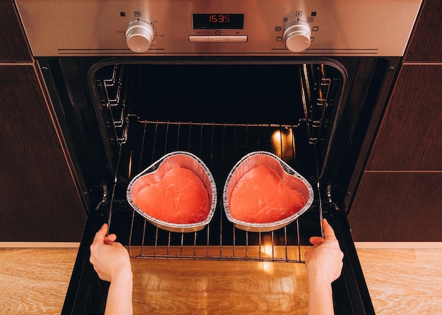 Een vrouw zet twee folievormen met rood deeg in de oven voor een hartvormige cake voor Valentijnsdag