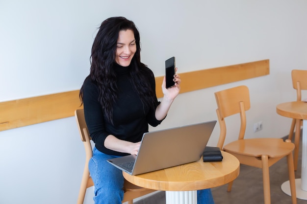 Een vrouw zit aan een tafel met een laptop en een telefoon in haar hand.