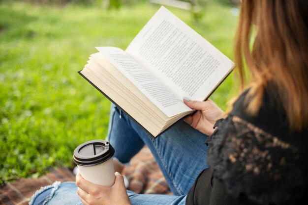 Een vrouw zit bij een boom in het park en houdt een boek en een kopje met een warme drank vast