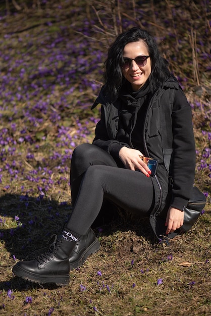 Een vrouw zit in het gras met haar benen over elkaar en haar been draagt een zwarte jas.