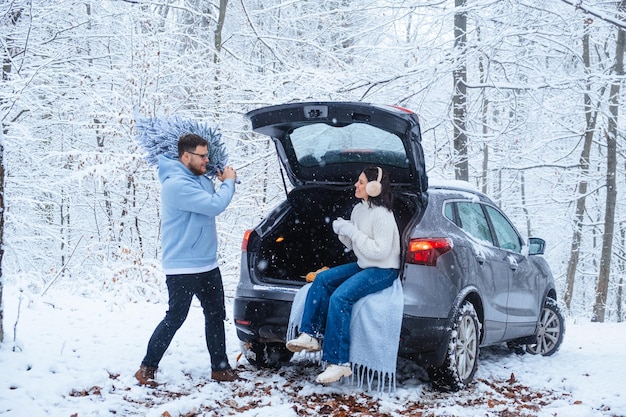 Een vrouw zit met een kopje in de kofferbak van een auto in een winterbos, een man draagt een kerstboom