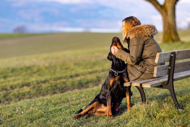 Een vrouw zit op een bank en knuffelt 's ochtends een rottweiler op een heuvel