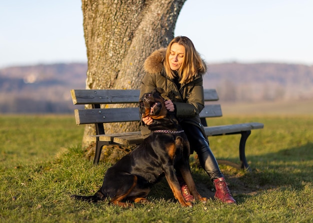 Een vrouw zit op een bank en knuffelt 's ochtends een rottweiler op een heuvel