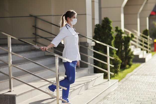 Een vrouwelijke arts of verpleegster die een beschermend masker en medische kleding draagt op de trappen van het ziekenhuis