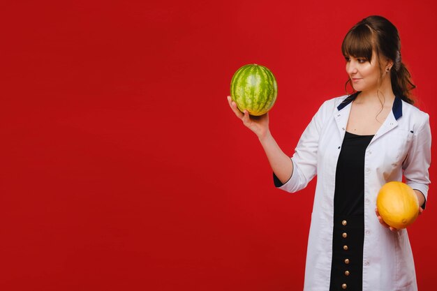 Een vrouwelijke arts-verpleegster in een witte jas met fruit in haar handen vormt op een rood