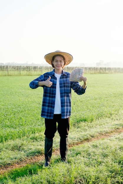 Een vrouwelijke Aziatische boer staat midden in een rijstveld met Thaise bankbiljetten en duim omhoog in het midden van een rijst veld bij zonsopgang