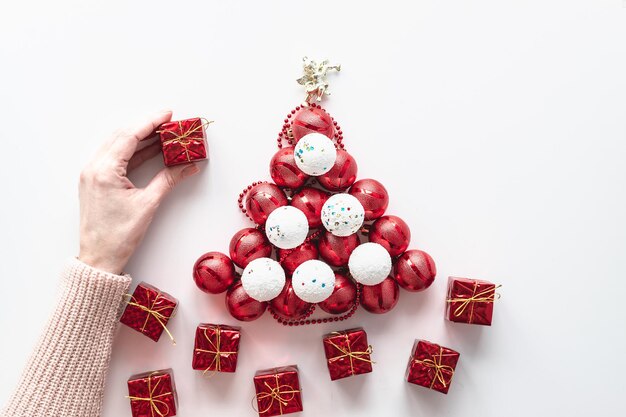 Een vrouwelijke hand houdt een kerstcadeau vast. Op een witte achtergrond, een kerstboom gemaakt van witte en rode ballen met geschenken. Feestelijke ansichtkaart.